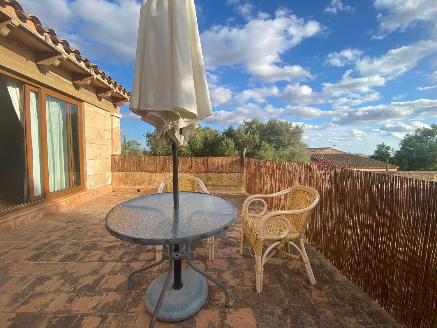 Sun-drenched terrace with bistro table and wicker chair under white umbrella against rustic backdrop.