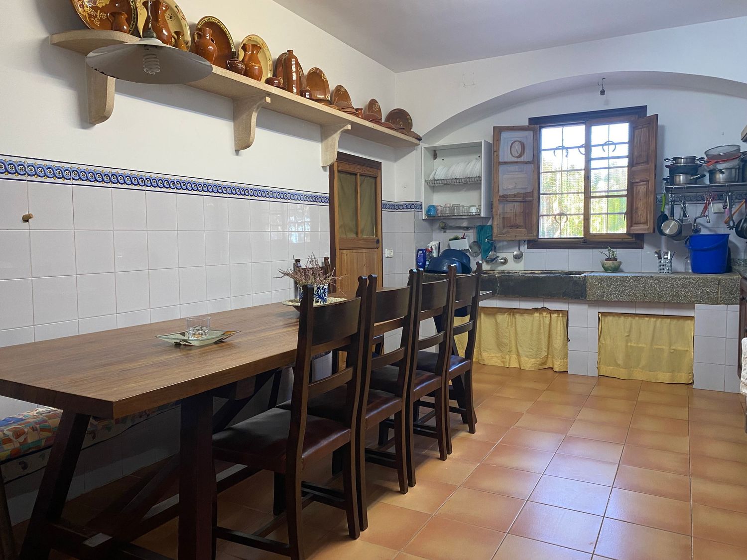 Rustic kitchen dining area with wooden table, chairs and decorative pottery displayed on wall shelves.