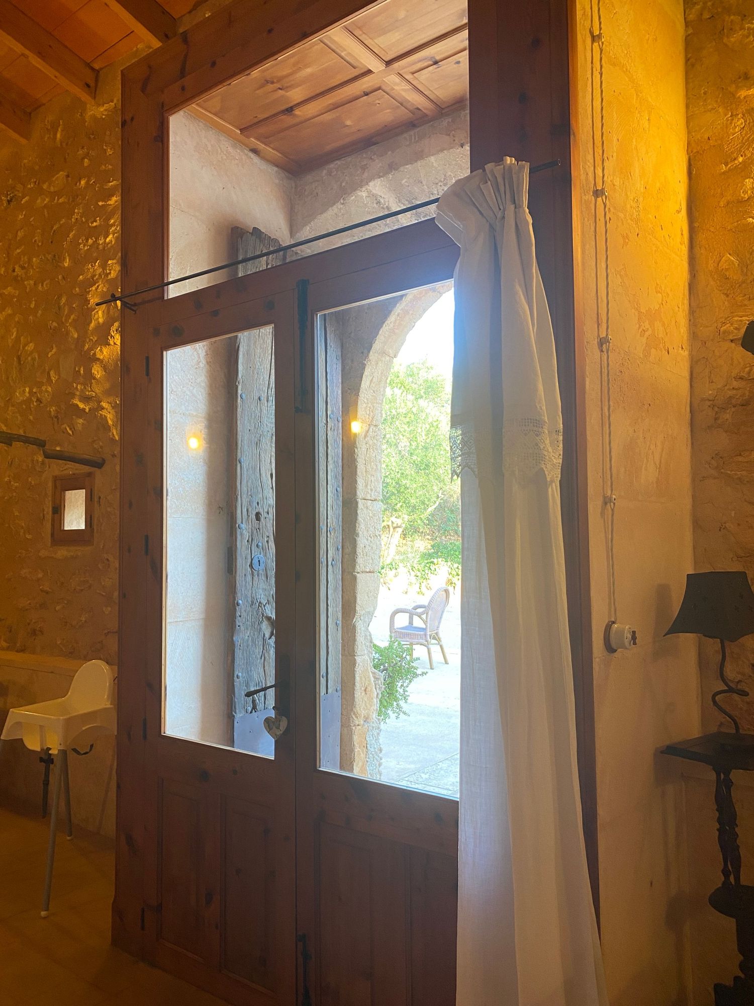 Large window with white curtains and wooden frame lets natural light into a rustic stone-walled interior.