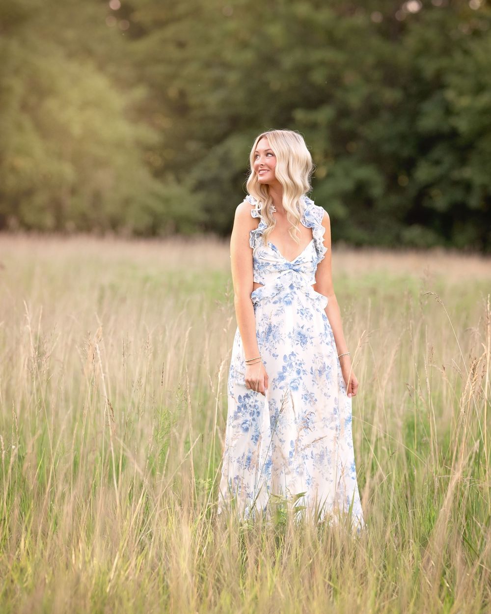 Woman in blue floral maxi dress standing in golden meadow with lush green trees in background. Kansas City senior portrait session -natural