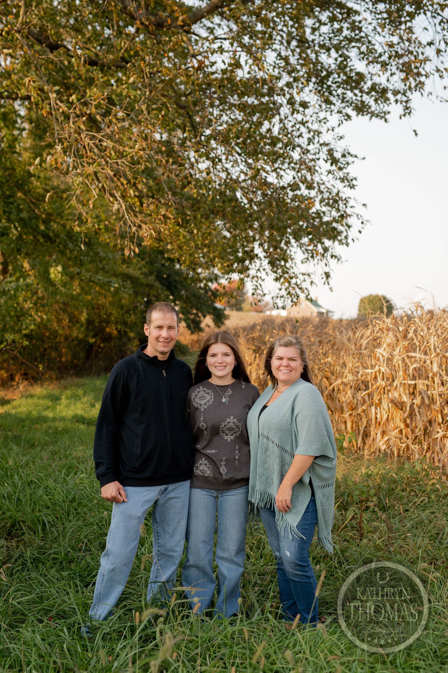 Family of three poses together in front of autumn cornfield at golden hour.