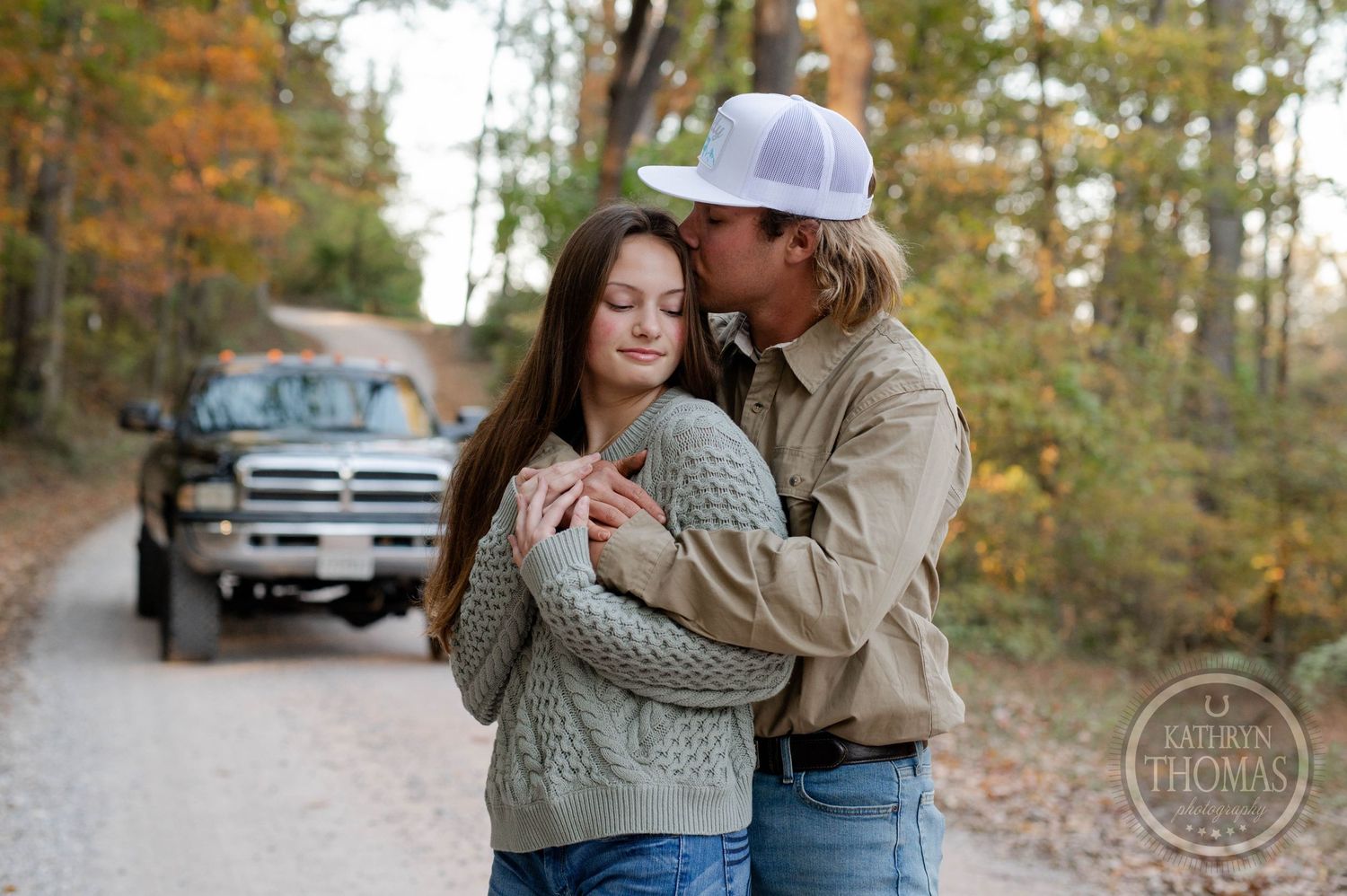 A couple shares an intimate moment on a fall forest path with a truck parked in the distance.