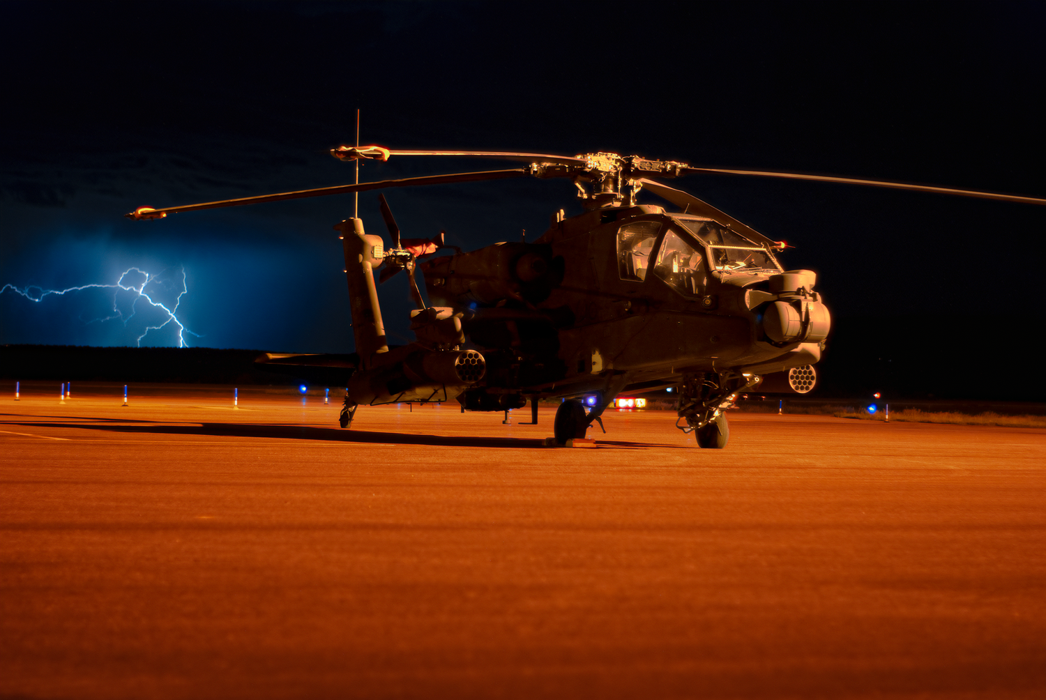 An AH-64 Apache helicopter sits on a dark airfield at night with lightning flashing in the background.