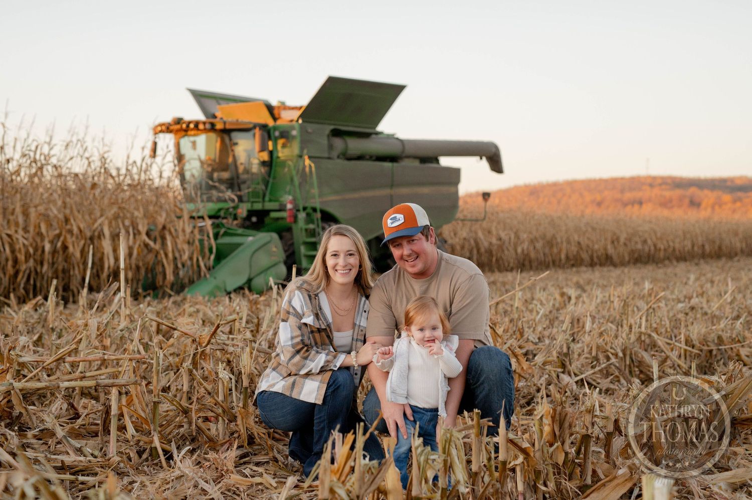 Family sits together in autumn cornfield with green John Deere combine harvester behind them.