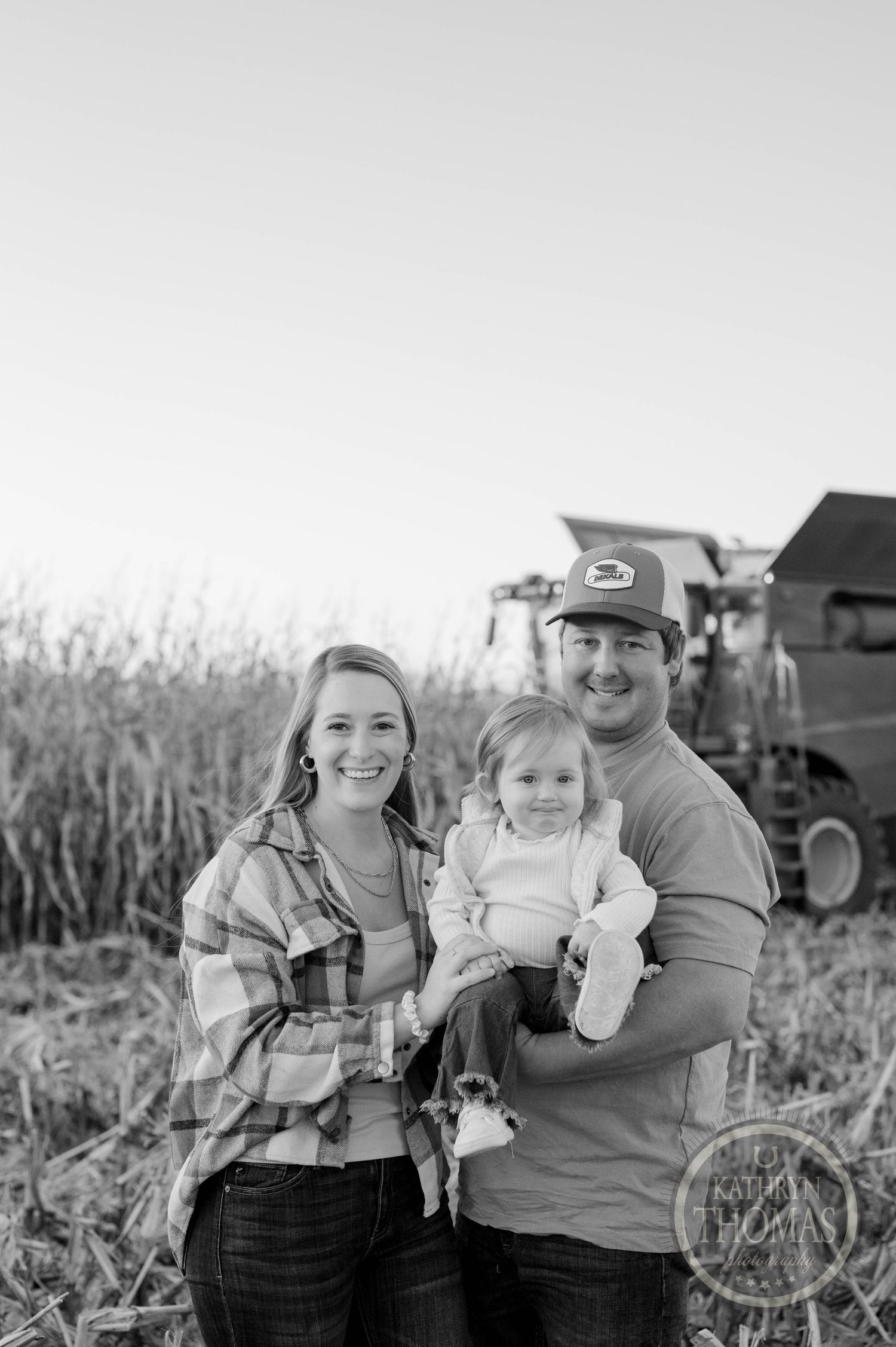 Black and white portrait of family standing in harvested cornfield with combine in background.