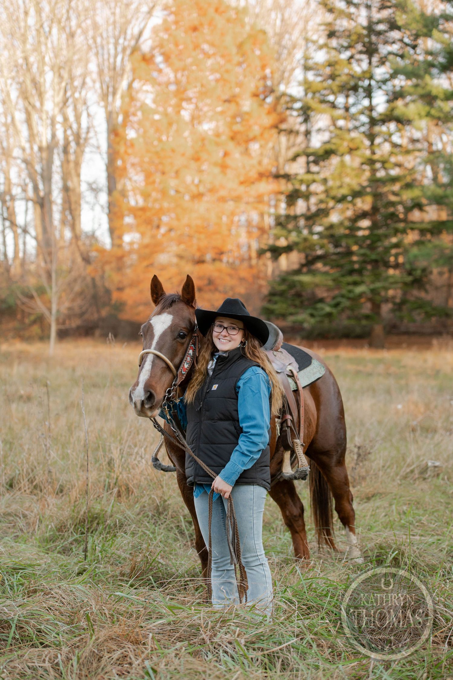 Horse and handler standing in autumn field with orange foliage and evergreen backdrop.
