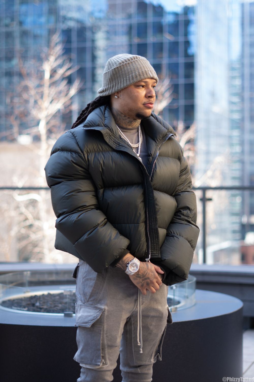 Person in warm winter attire with beanie and puffy jacket poses against urban backdrop of glass buildings.