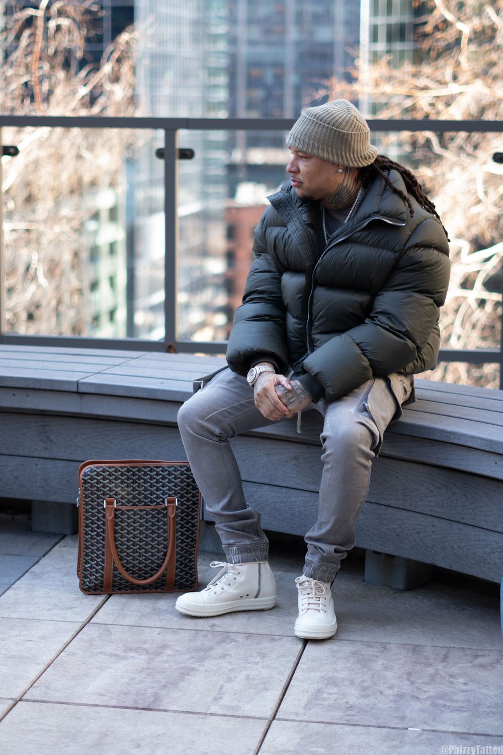 Person wearing puffy black coat and grey sweatpants sits with designer bag on urban terrace.