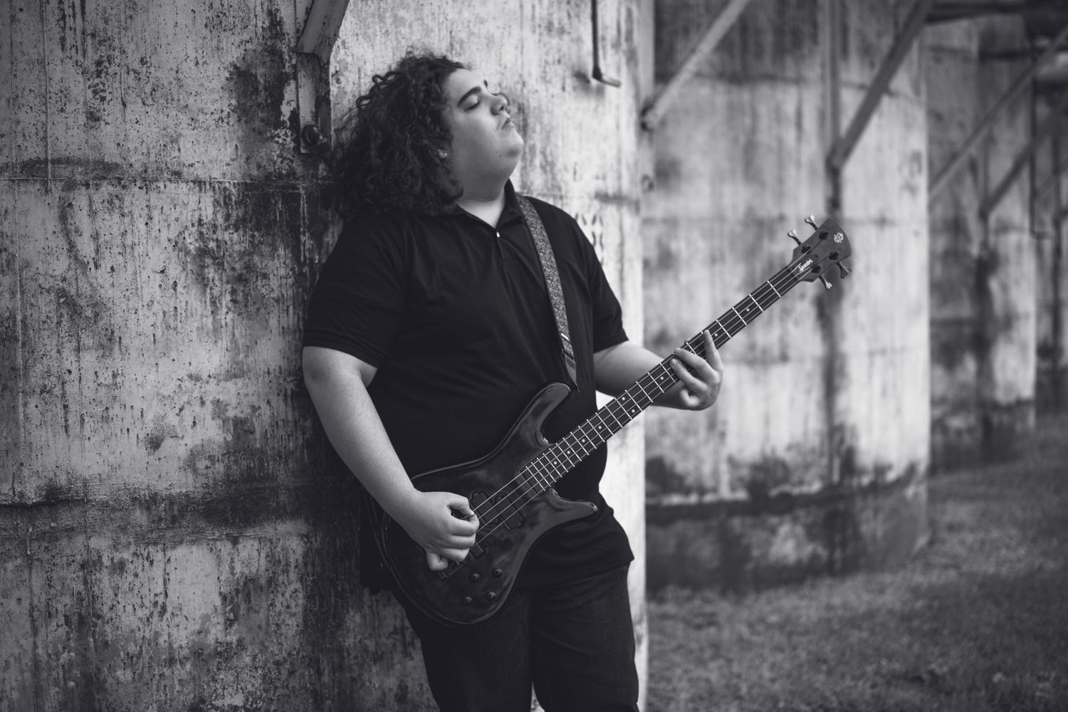 A musician in black clothing plays guitar against a textured wall in dramatic black and white lighting.