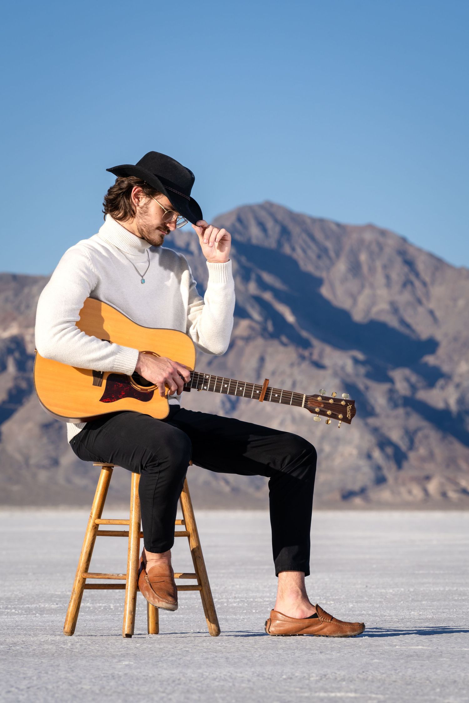 A musician in a cowboy hat playing acoustic guitar while sitting on a stool in a desert landscape.