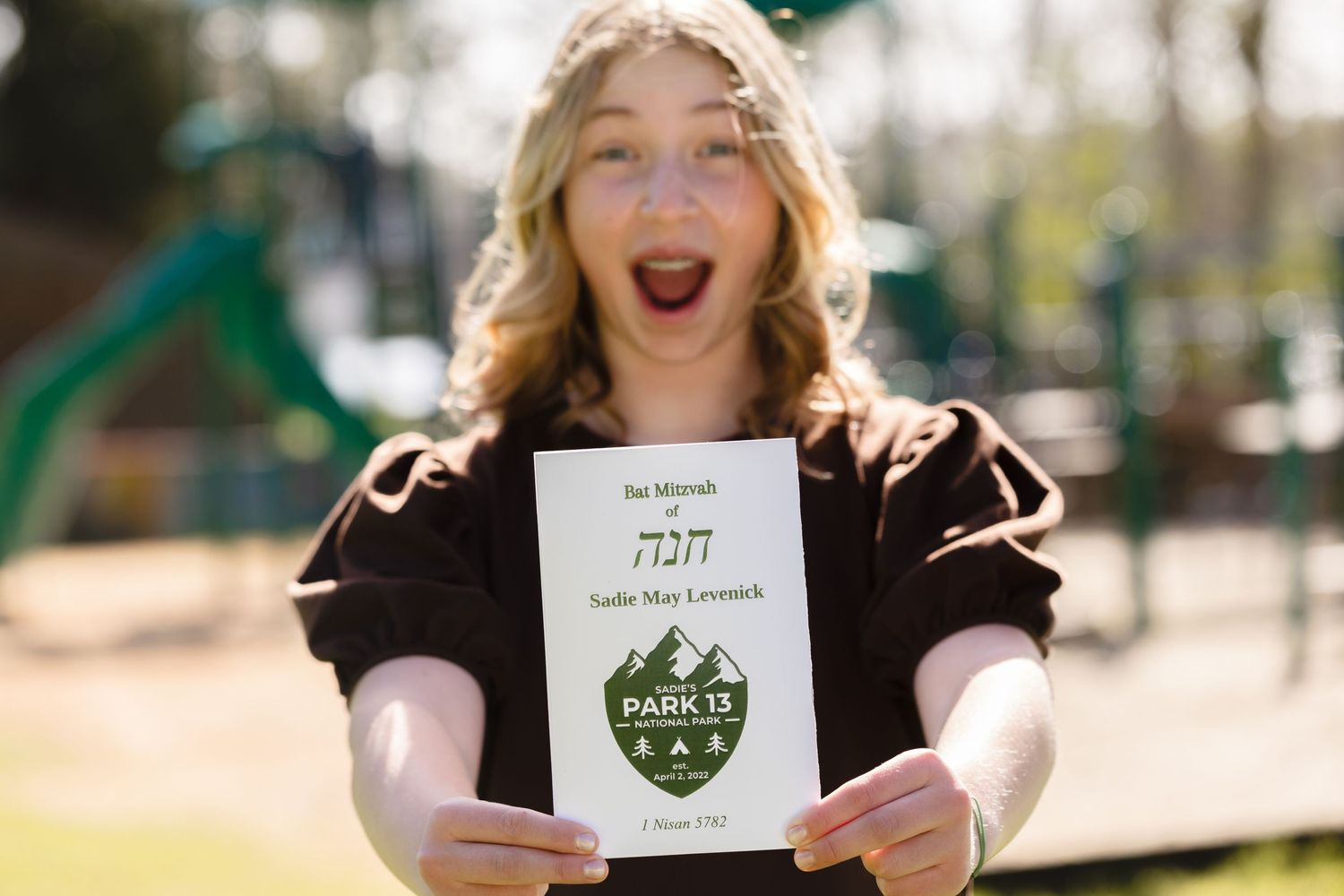 An excited looking person holds up a card with Hebrew text and a green crest design in bright outdoor lighting.