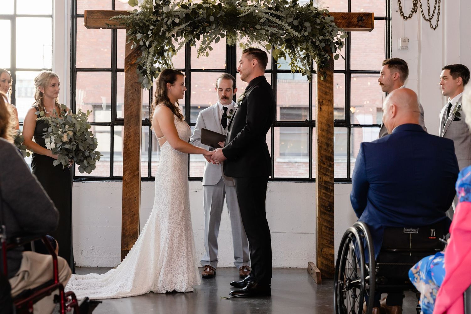 Wedding ceremony taking place under a greenery arch in an indoor venue with guests in attendance.
