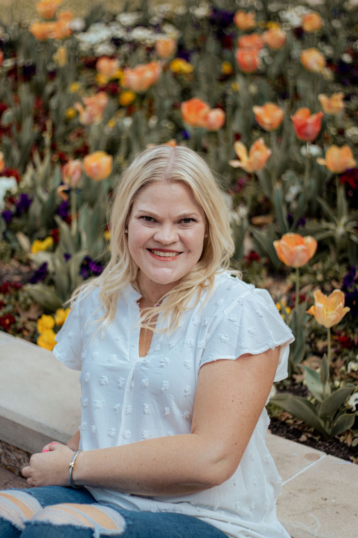 Woman in white eyelet top sits in front of colorful garden with orange and yellow tulips blooming in background.