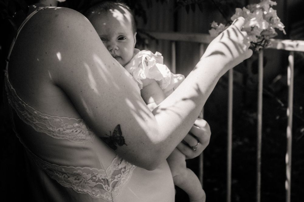Parent in lace dress holding baby outdoors with subtle black and white tones.