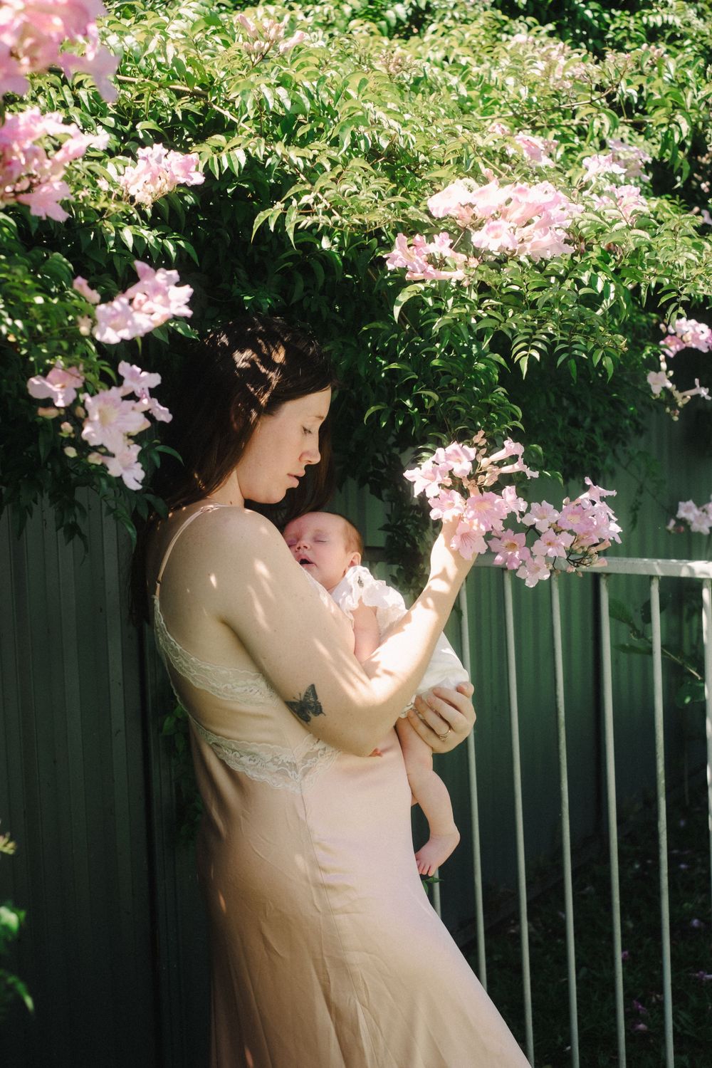 Intimate maternal moment under blooming pink flowers against a green garden fence.