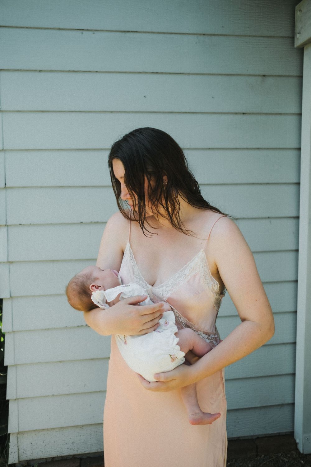 Person in peach dress holding baby against white siding.