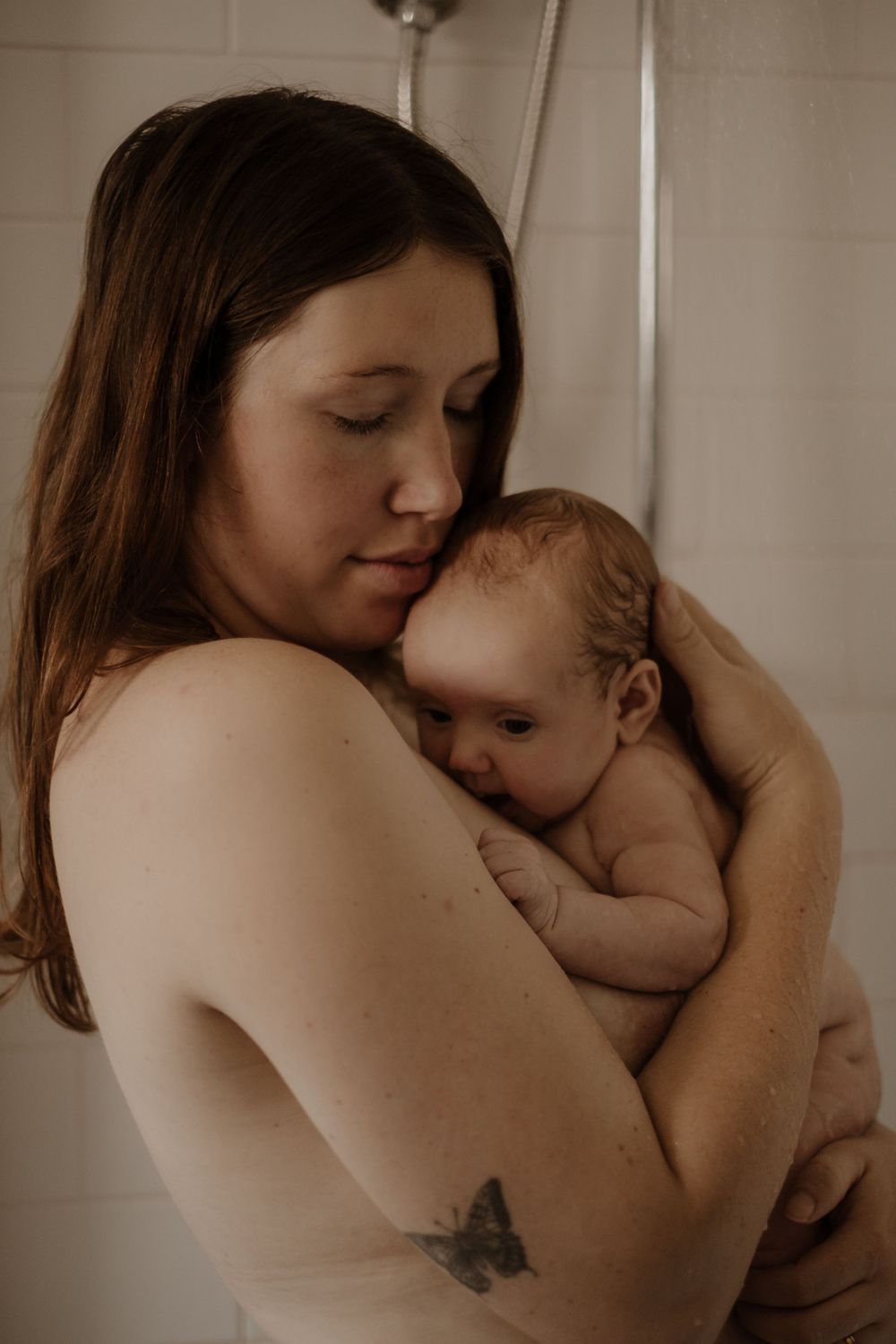 A tender moment between parent and newborn baby showing skin-to-skin contact and a butterfly tattoo in muted tones.