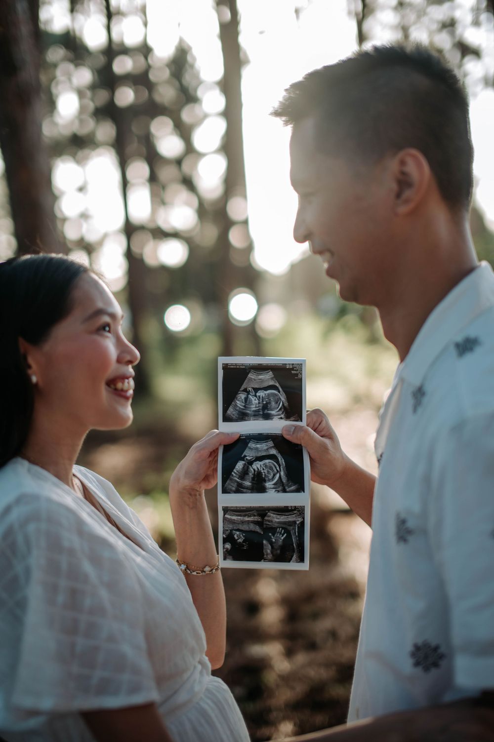 Expectant couple sharing ultrasound photos in a sunlit forest setting, displaying their joy and anticipation.