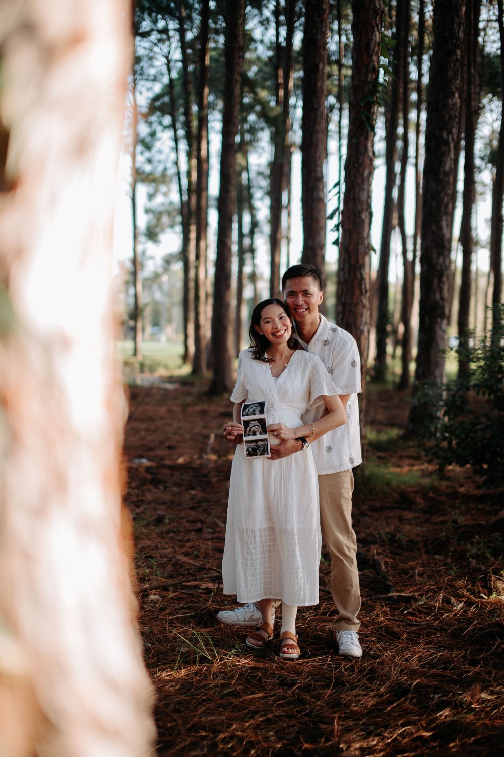 Smiling couple in white outfits pose with ultrasound photos in a forest setting for pregnancy announcement.