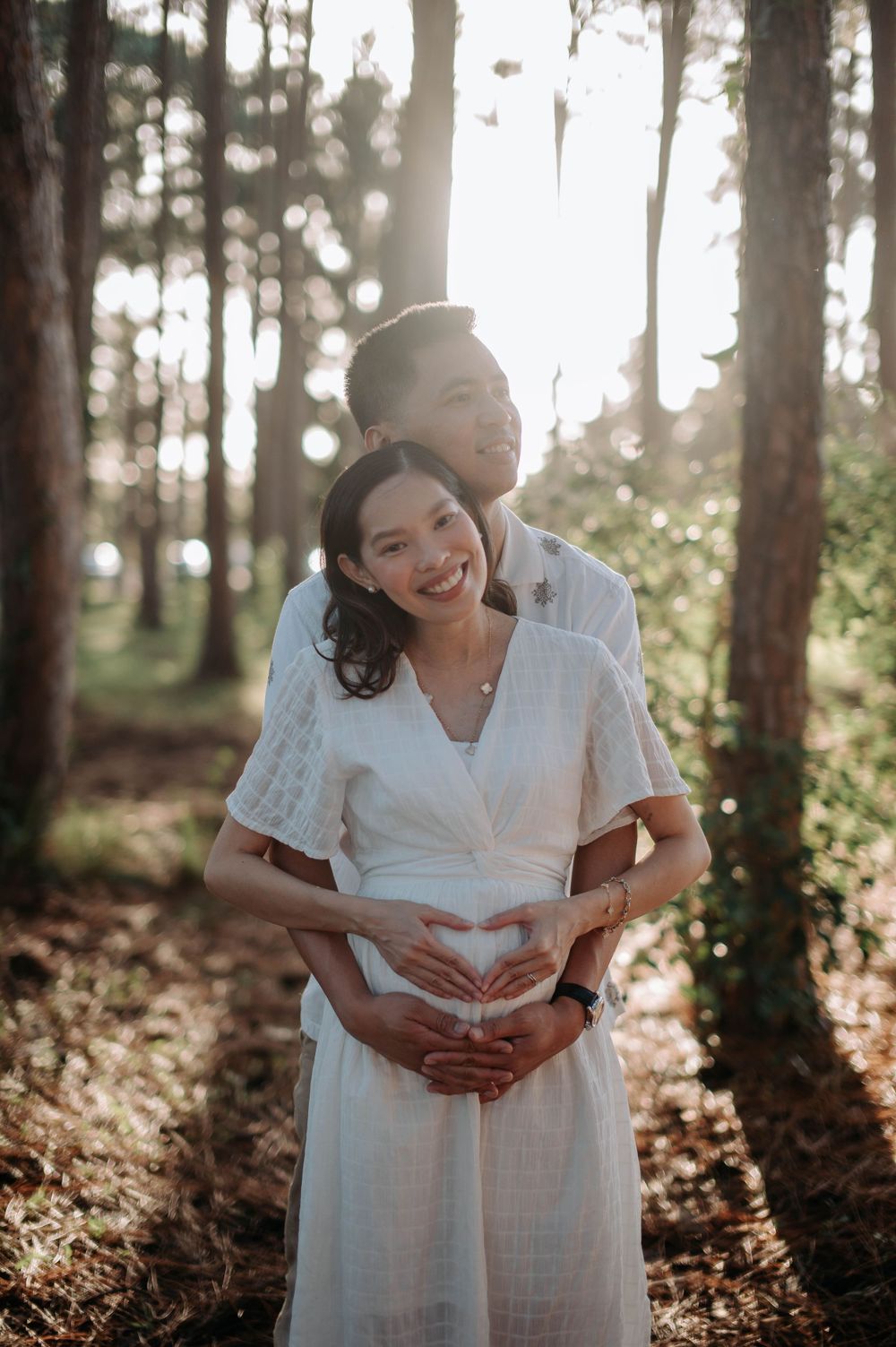 Happy couple embrace in forest while making heart shape with hands on pregnant belly.