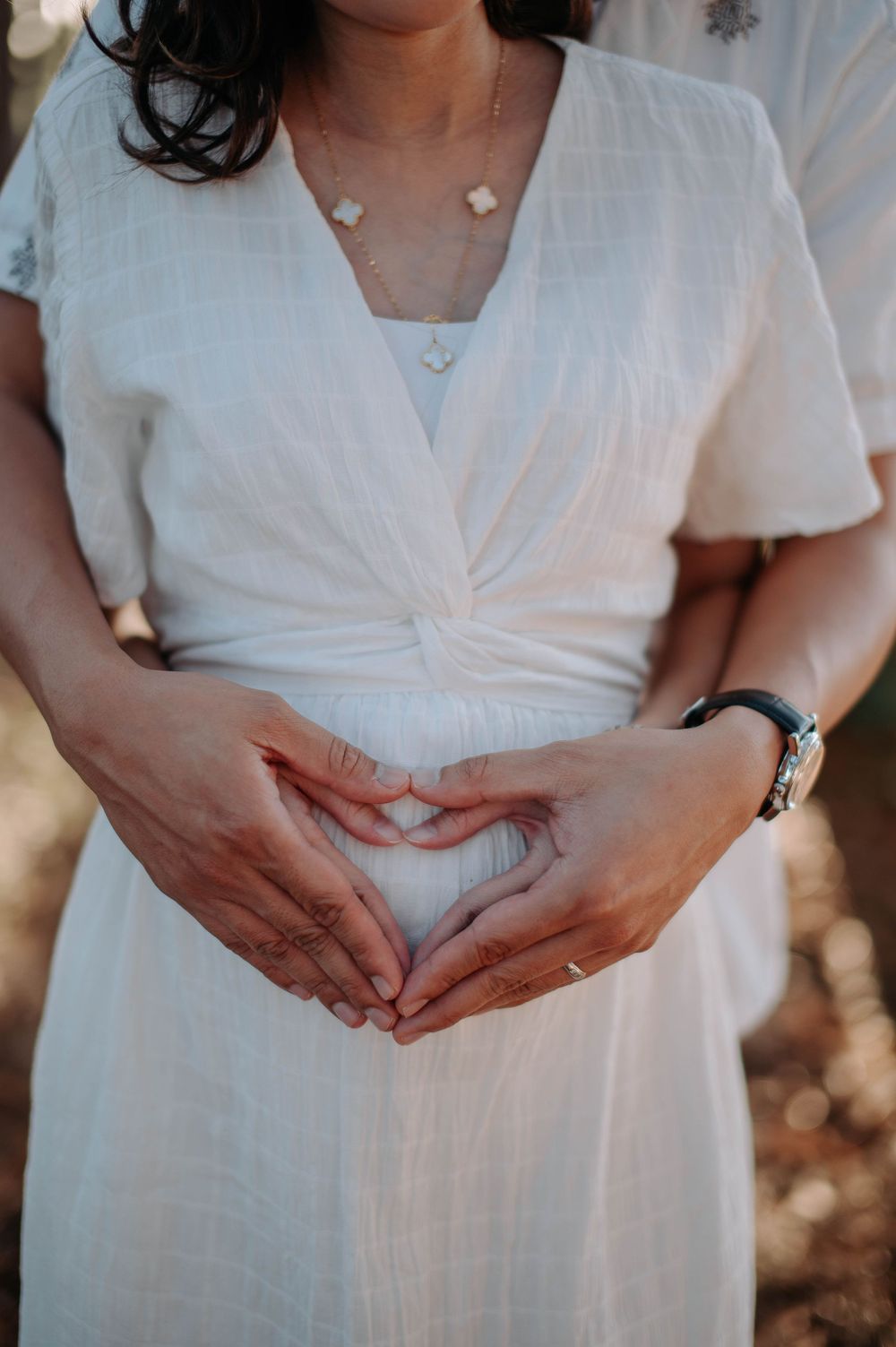 Close-up of hands forming heart shape on white maternity dress in forest setting.