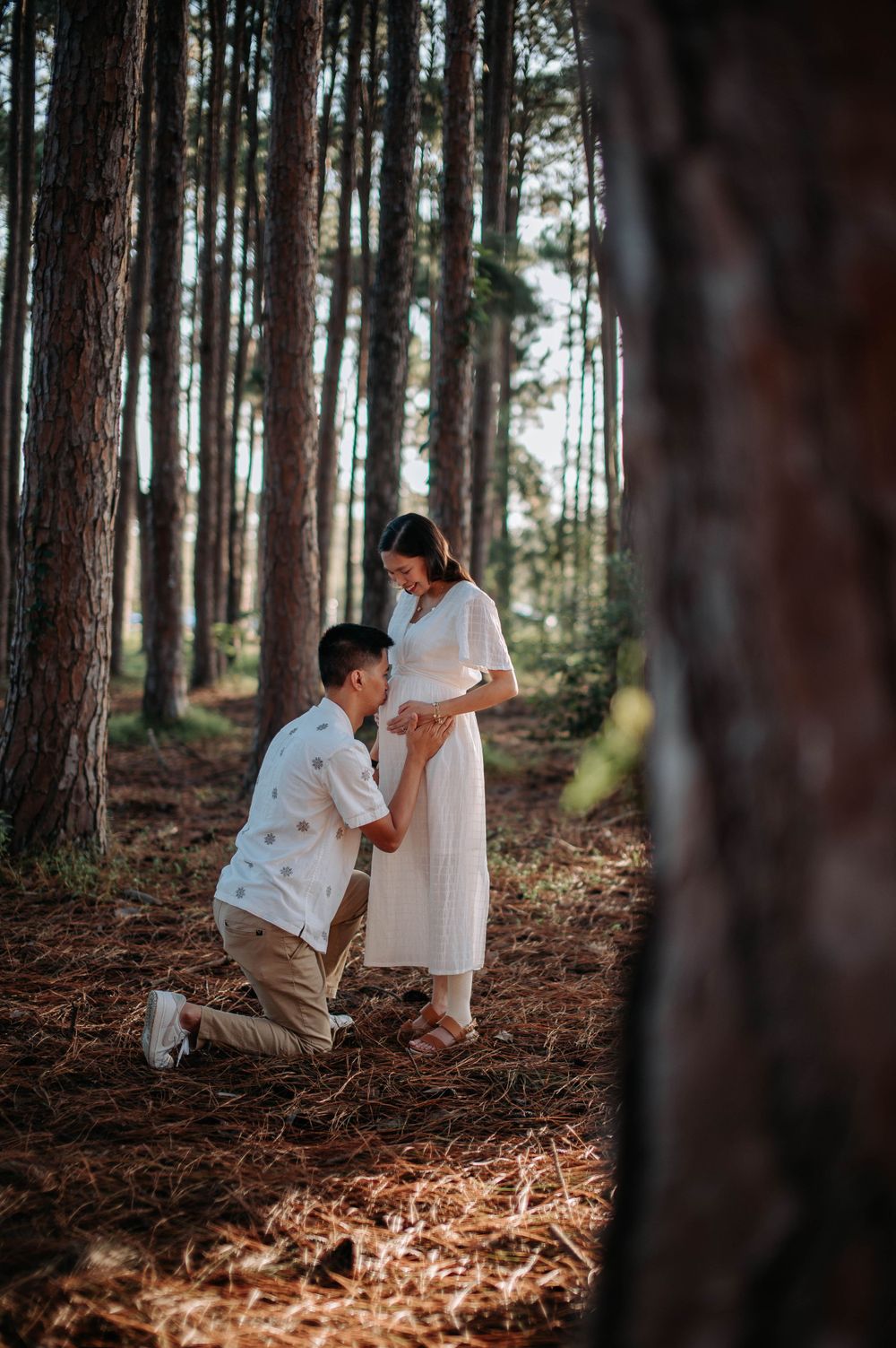 Partner kneels before pregnant woman in white dress during forest photoshoot.