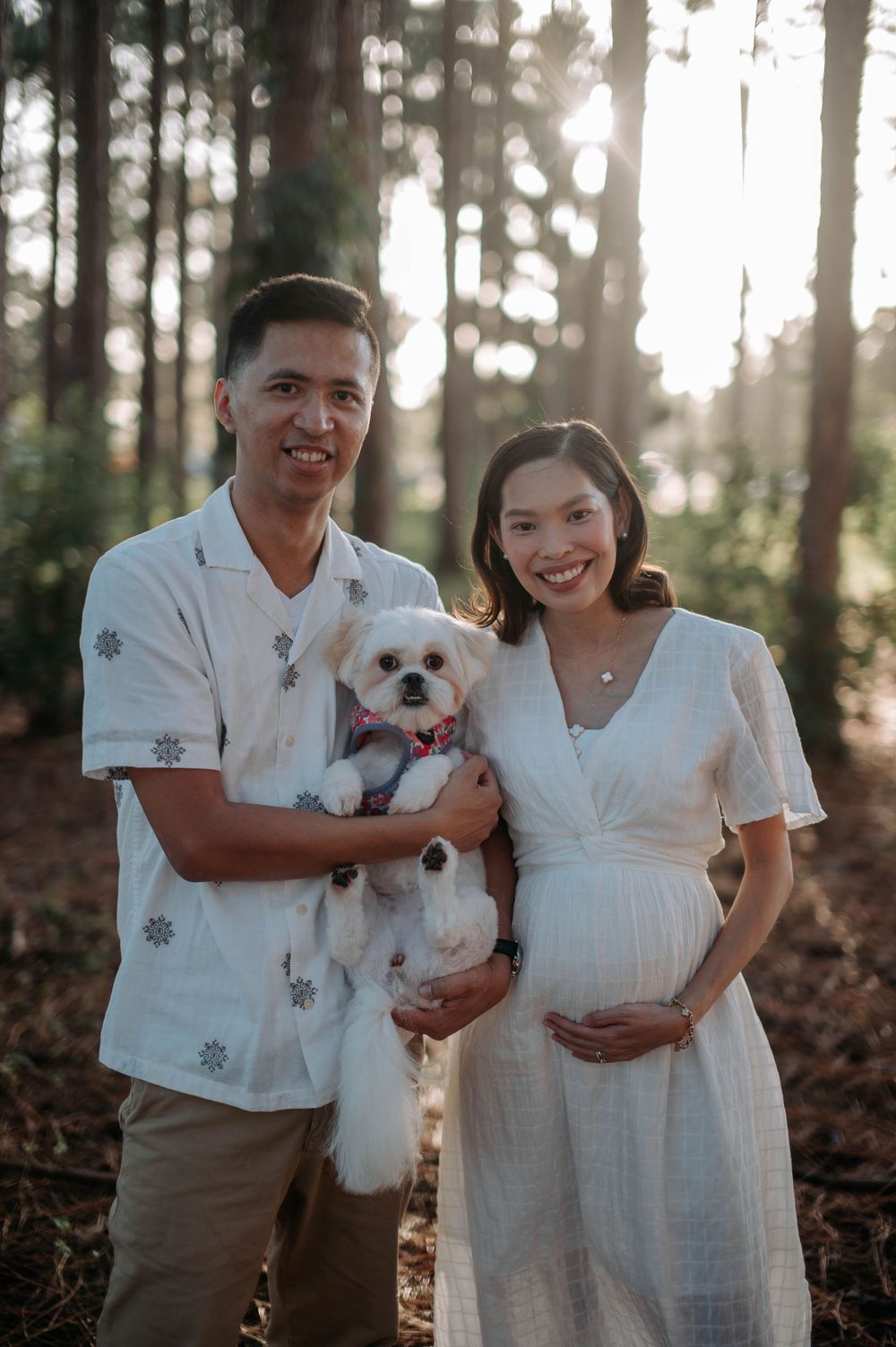 Expecting couple with small white dog pose for maternity photos in pine forest.