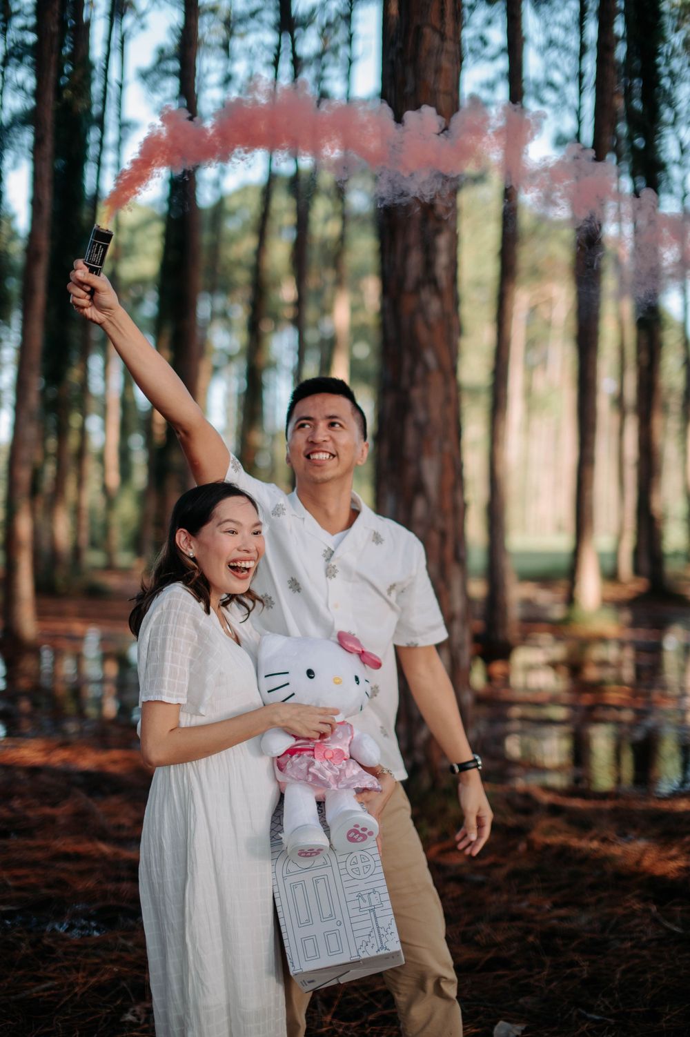 Couple celebrating with pink smoke bombs and a Hello Kitty plush in a wooded area.
