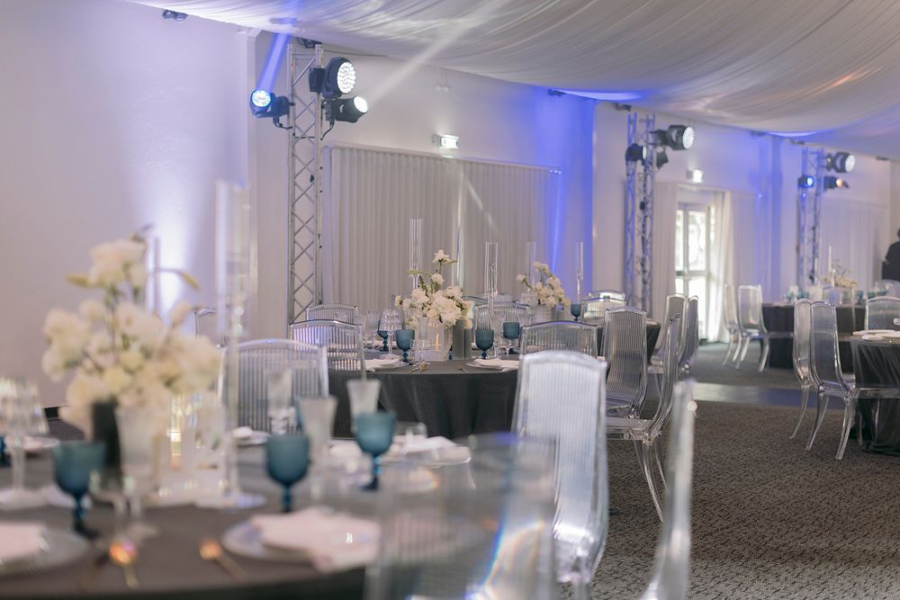 Elegant wedding reception venue with blue uplighting, white floral centerpieces, and ghost chairs.