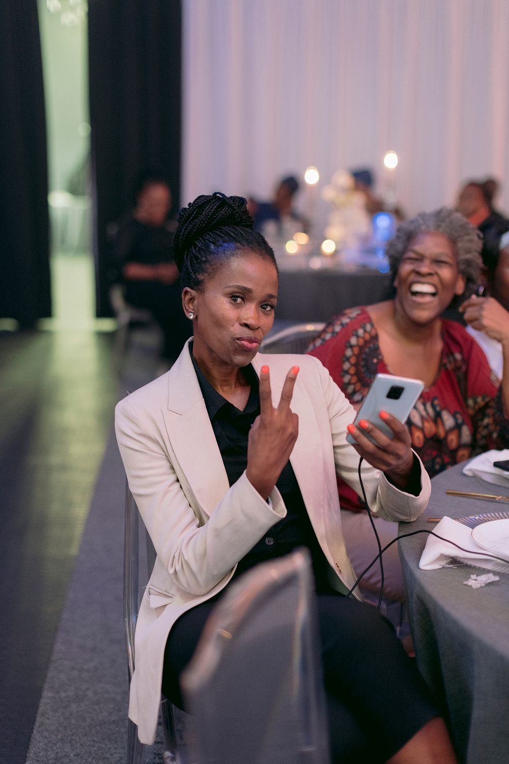 Woman giving peace sign while holding phone at formal banquet event.