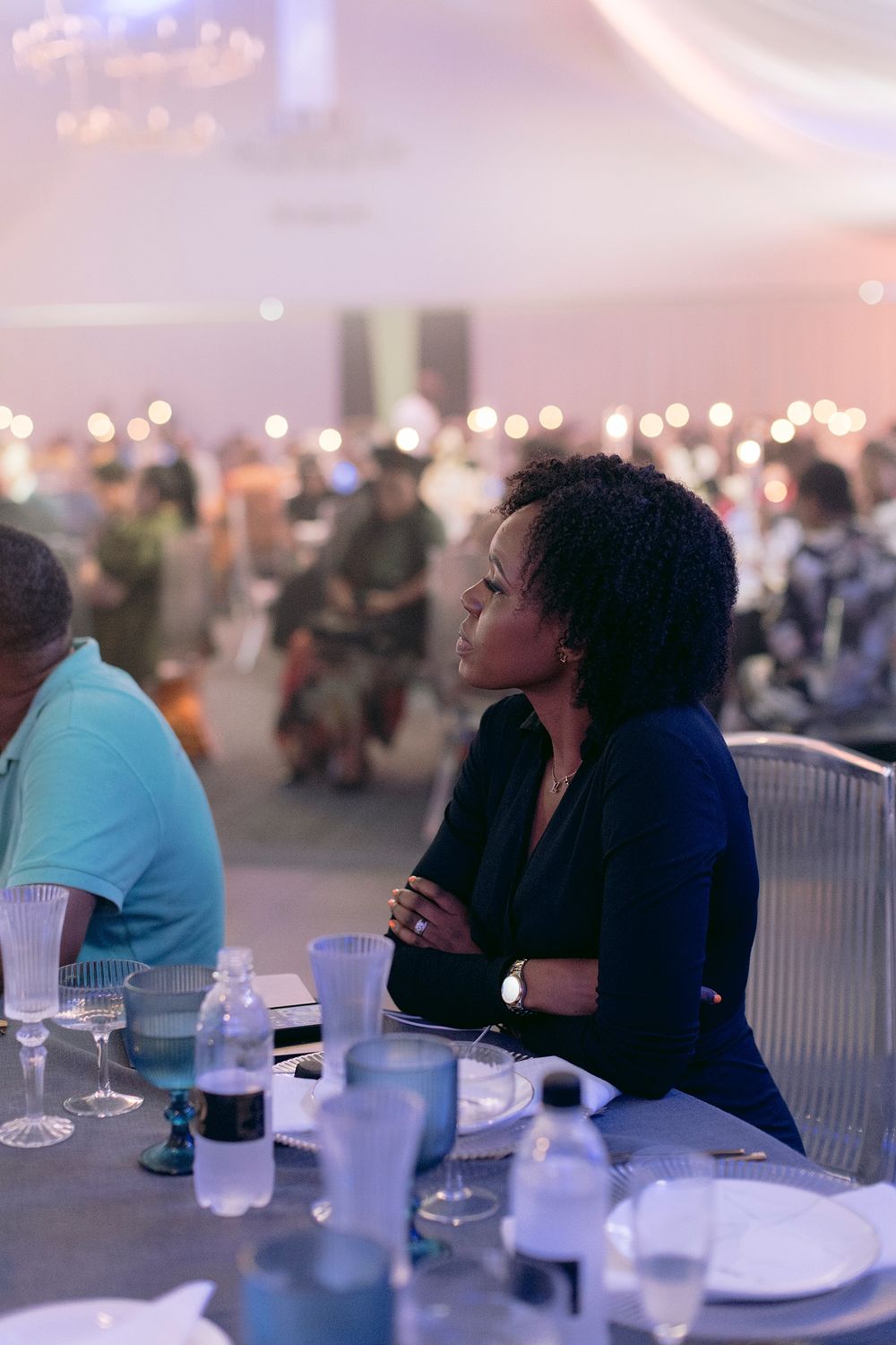 Woman in navy blazer sitting attentively at gala dinner table with arms crossed.