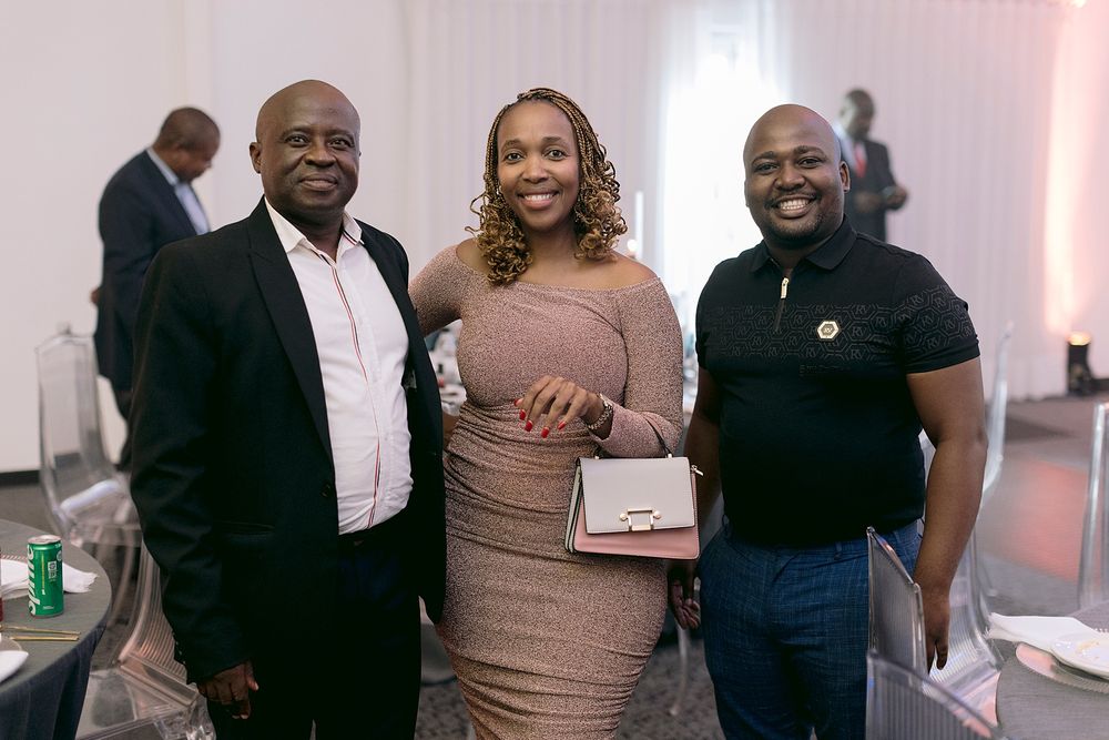 Three people smiling together at an indoor event, woman in center holding a white handbag.