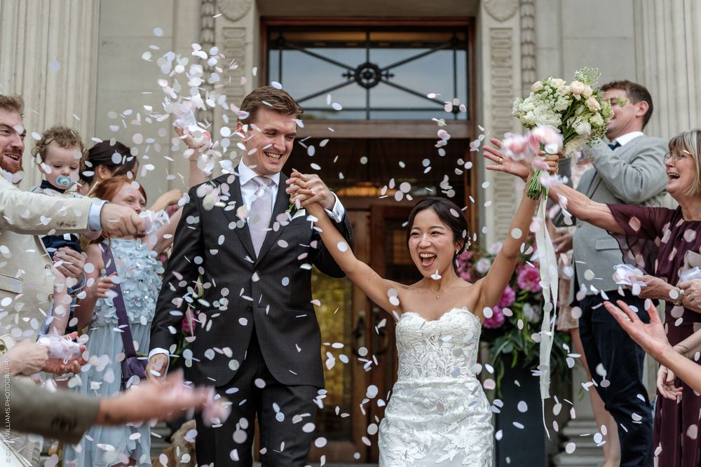 Joyful newlyweds exit wedding venue as guests shower them with pink and white confetti petals.