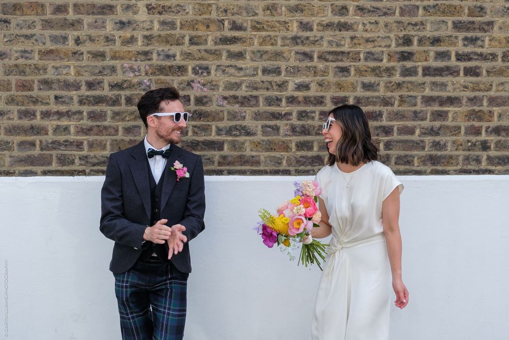A stylish wedding couple in sunglasses pose against a brick wall, the bride holding a vibrant colorful bouquet.