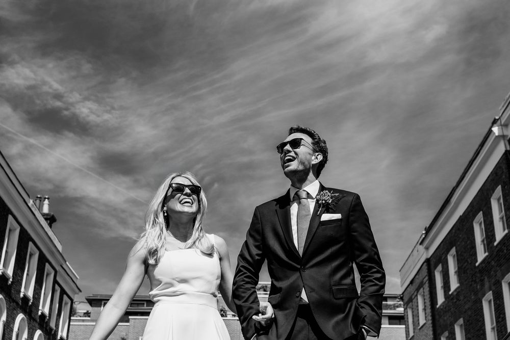 Black and white photo of a smiling couple in wedding attire holding hands under a dramatic cloudy sky.