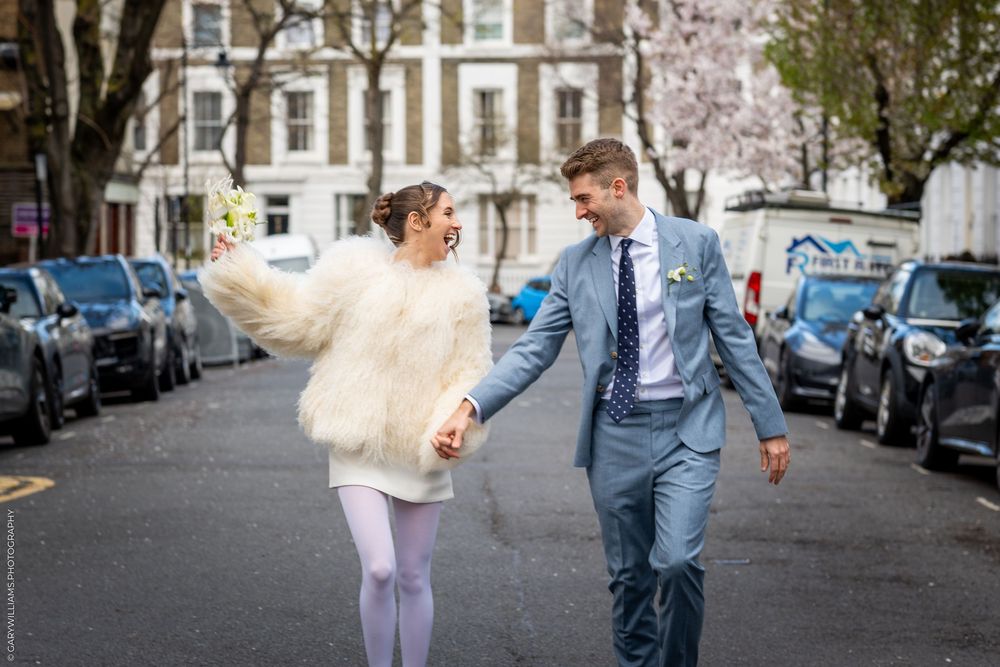 A couple runs playfully down an Islington street lined with parked cars and blooming cherry trees on a spring day.