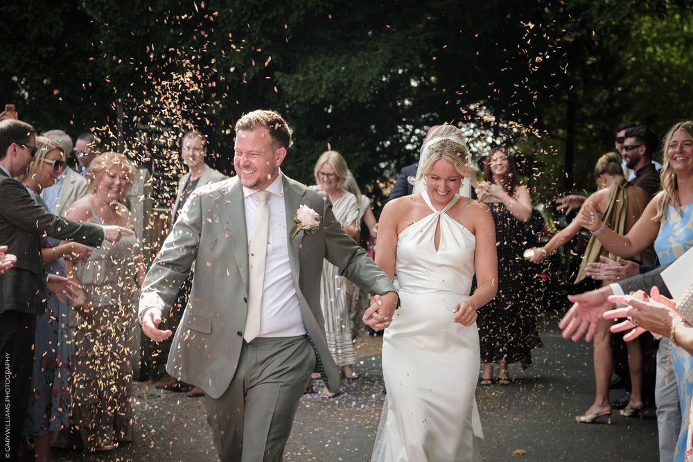 Bride and groom walk through confetti shower surrounded by cheering wedding guests outdoors.