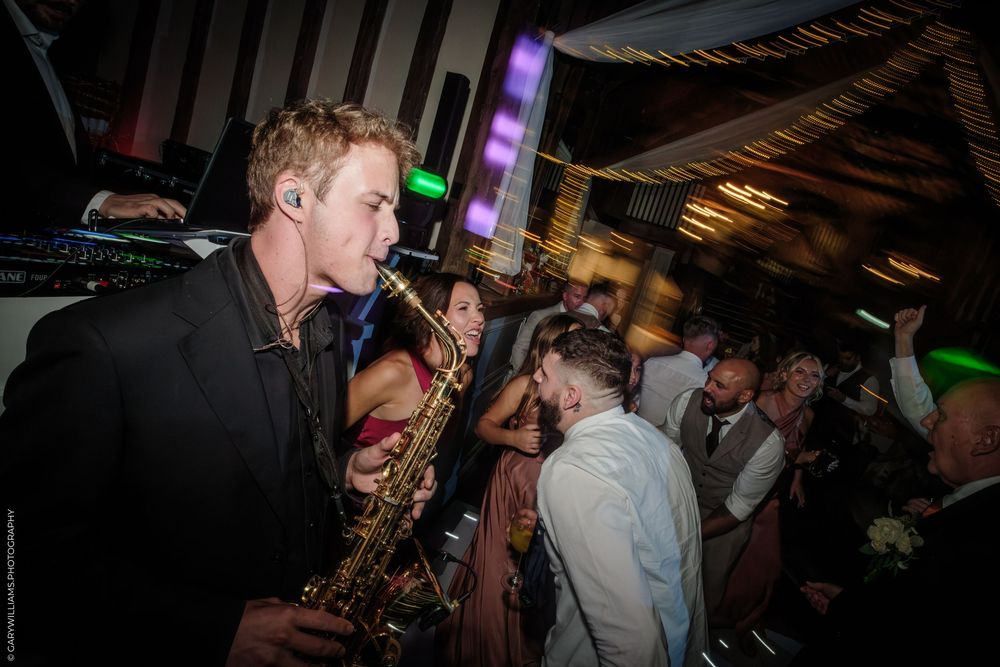 Saxophonist in black suit plays at lively wedding reception with dancing guests and purple lights.