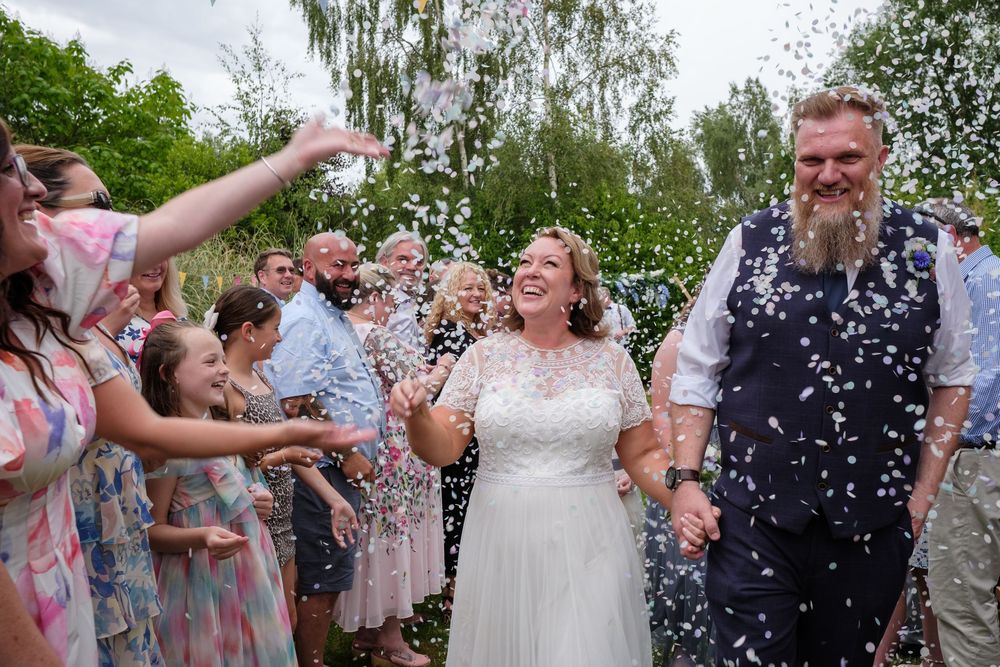 Wedding guests shower the happy couple with white confetti during an outdoor celebration in a lush garden setting.