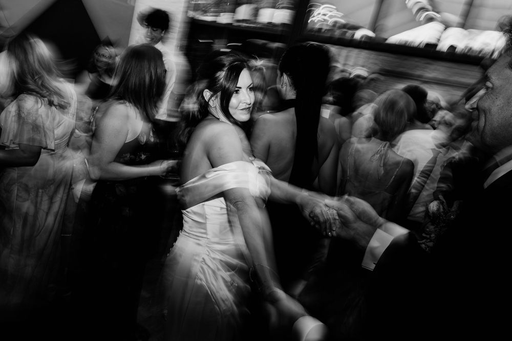 Black and white photo of a bride dancing energetically at her wedding reception surrounded by guests.