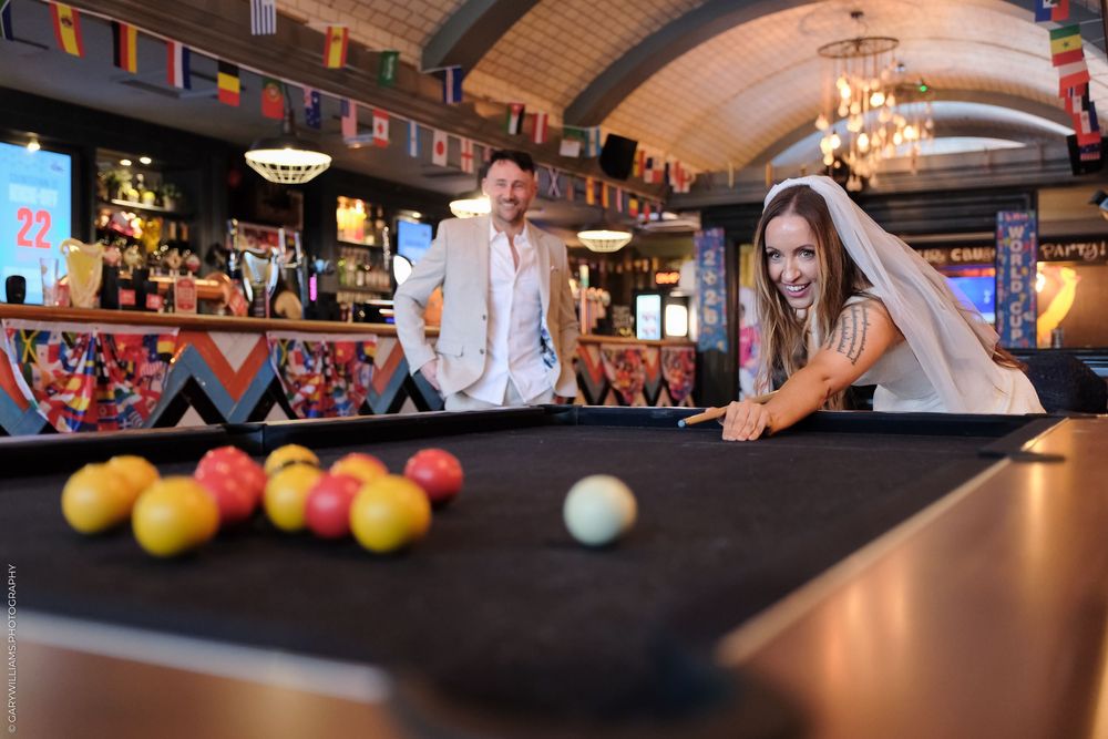 Bride in wedding veil plays pool at a bar while groom watches and smiles in a colorful venue.