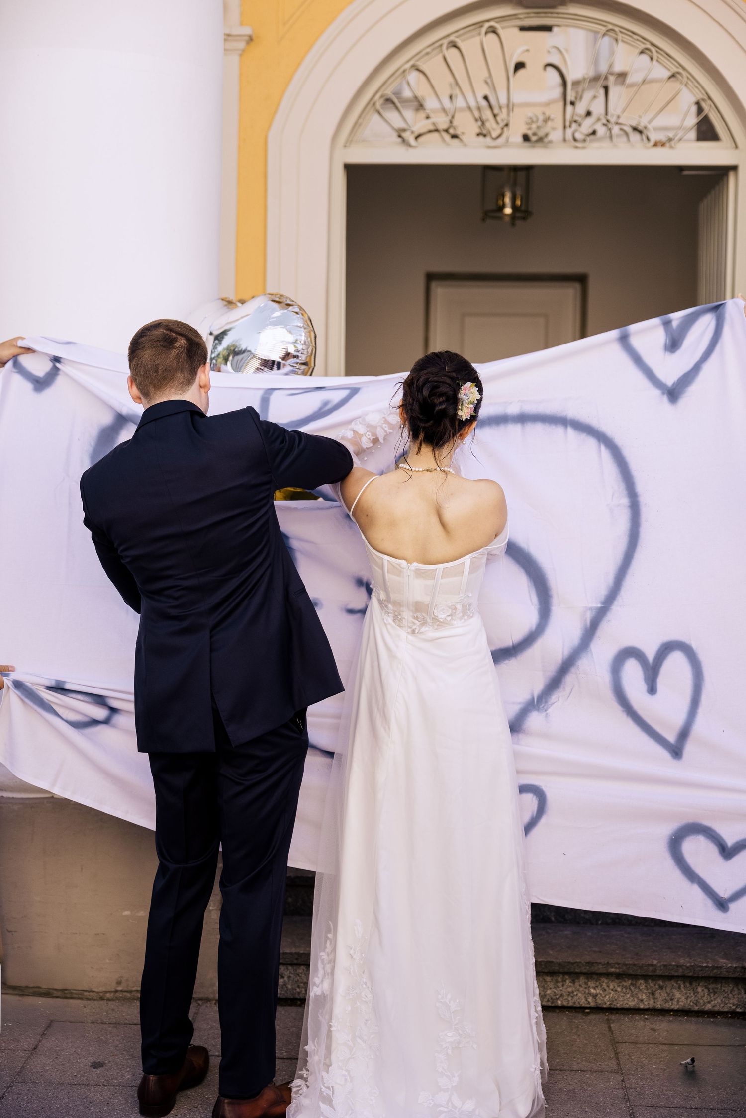 Newlyweds in formal attire hang decorative banner outside yellow building with arched entrance.