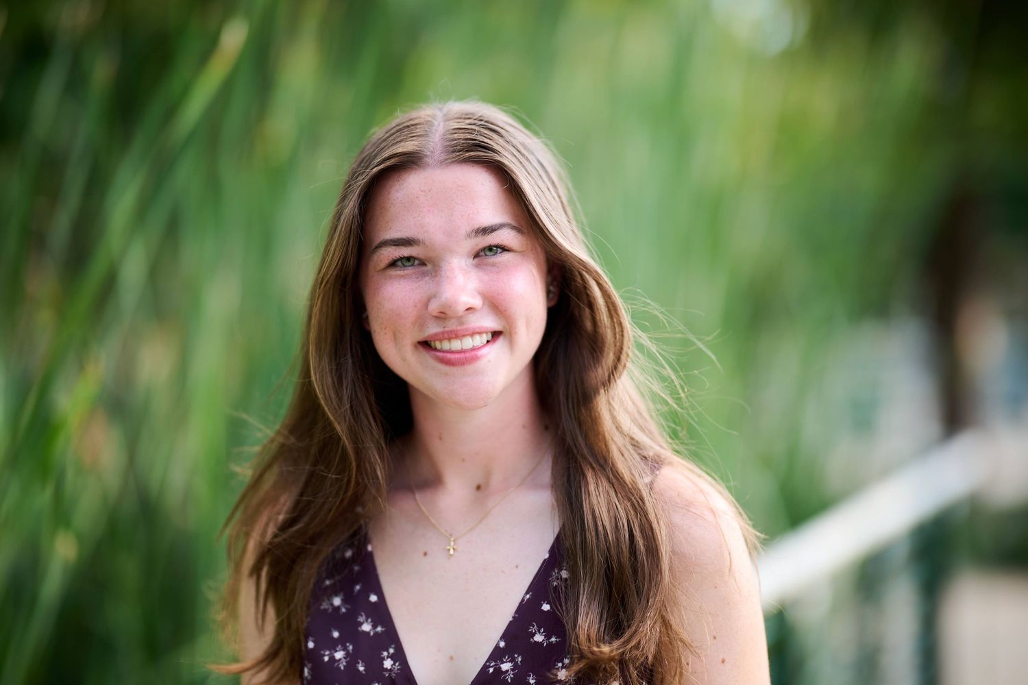 A young woman in a purple top smiles warmly while standing in front of green foliage.