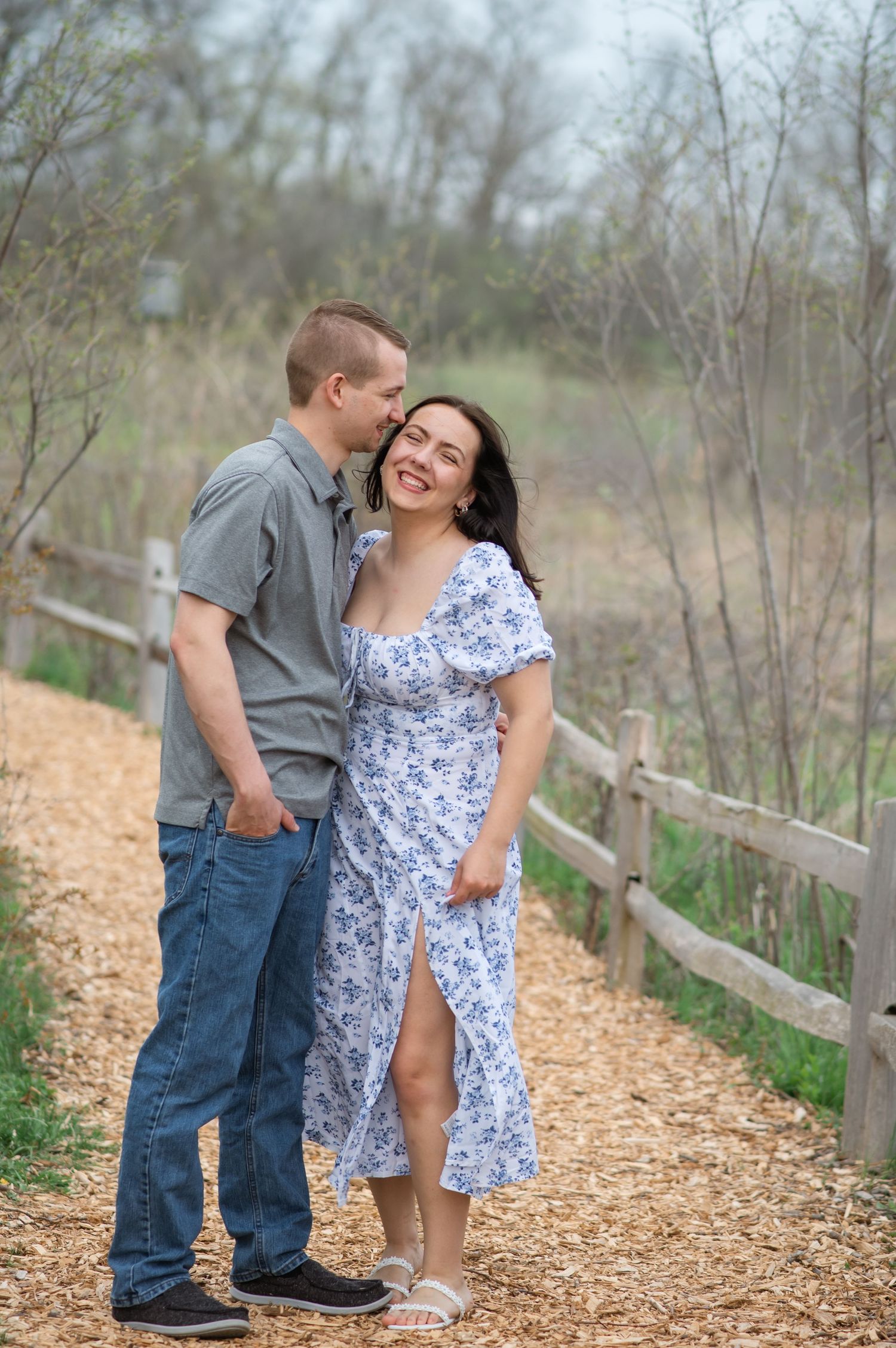 A couple shares an intimate moment on a wood chip path with a rustic fence and spring foliage in the background.