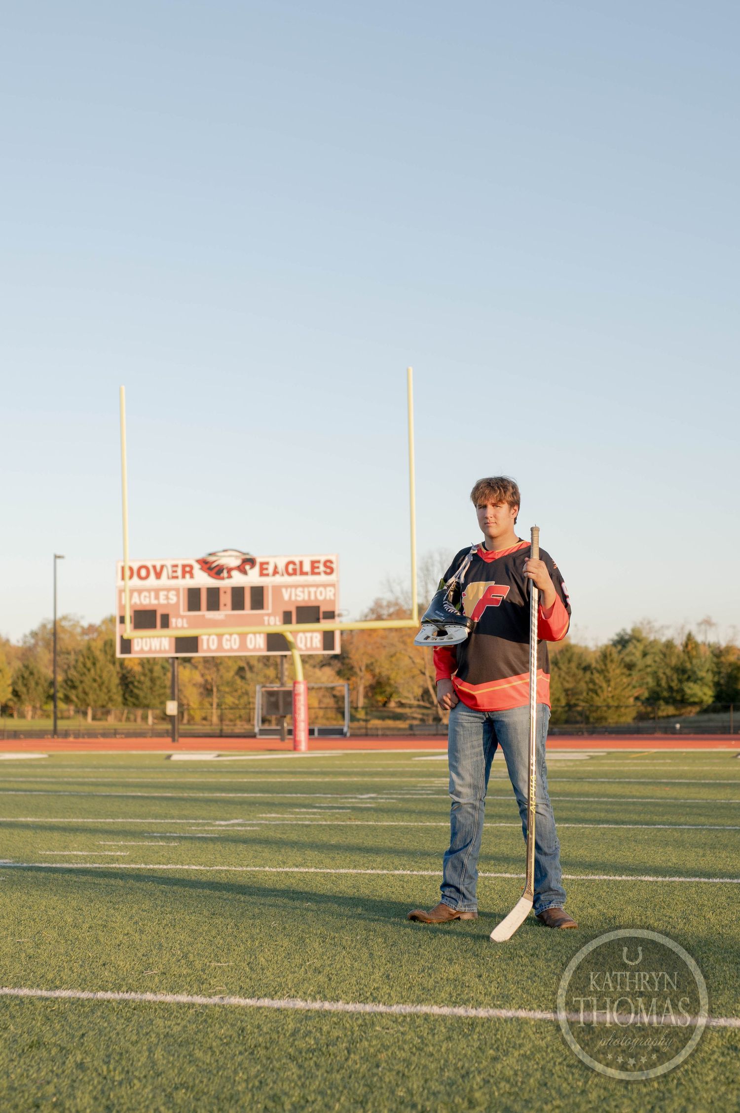 Student athlete stands with hockey stick and gear on football field at Dover Eagles stadium.
