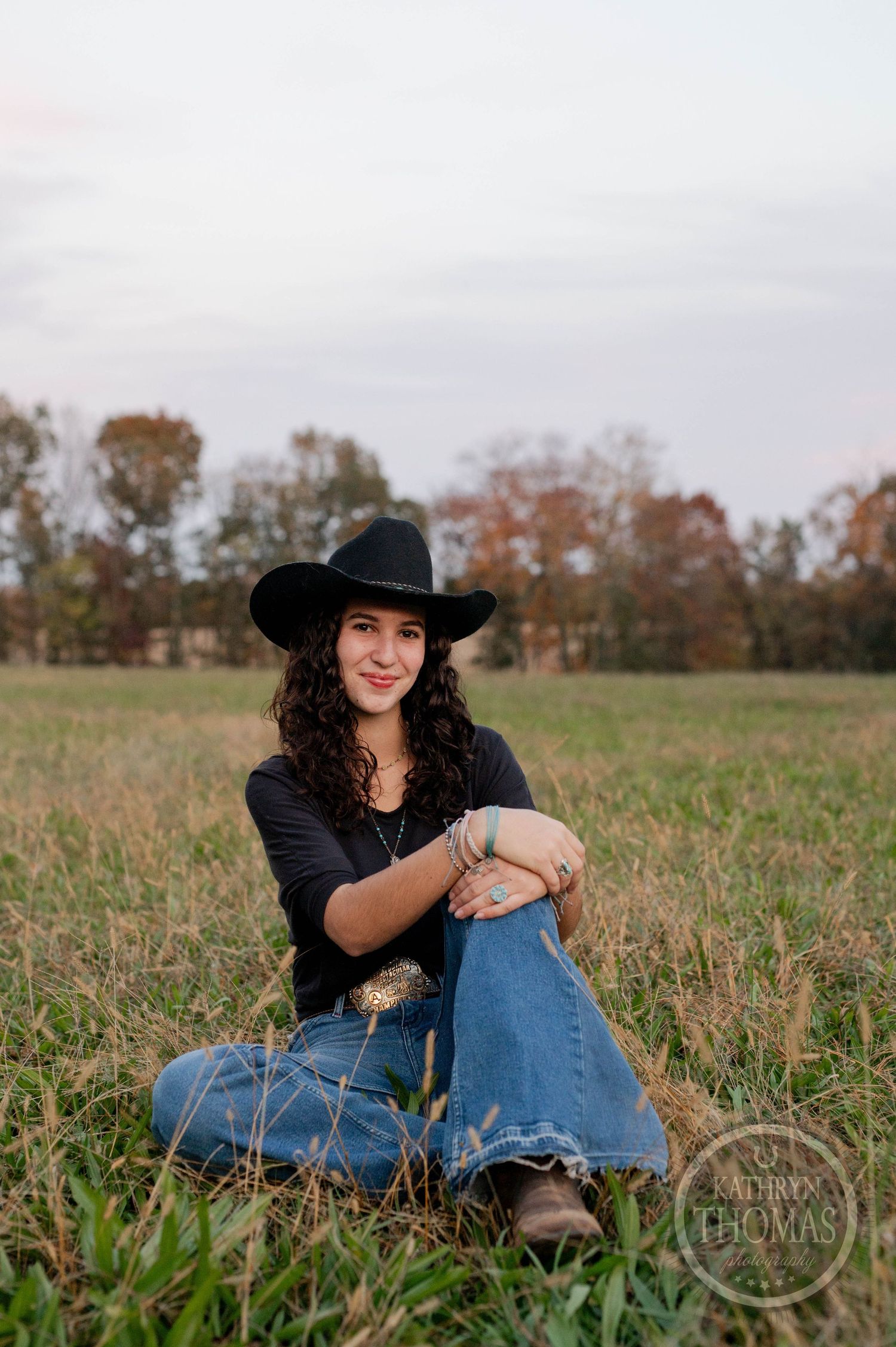 Casual portrait of person in western wear sitting in rustic field at sunset.