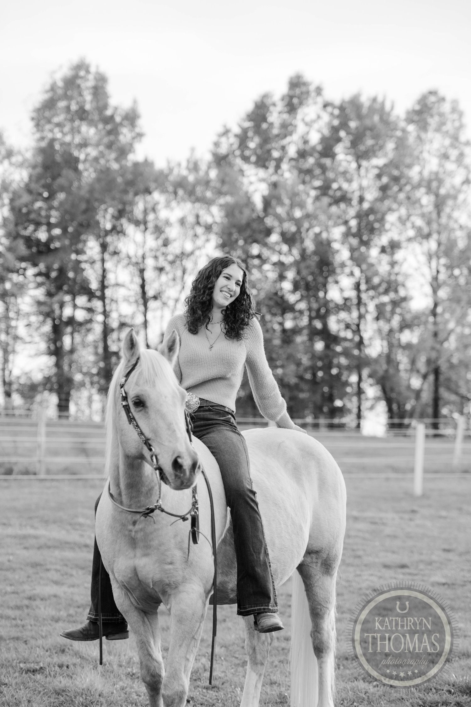 A black and white portrait of someone sitting on a pale horse in front of tall trees.