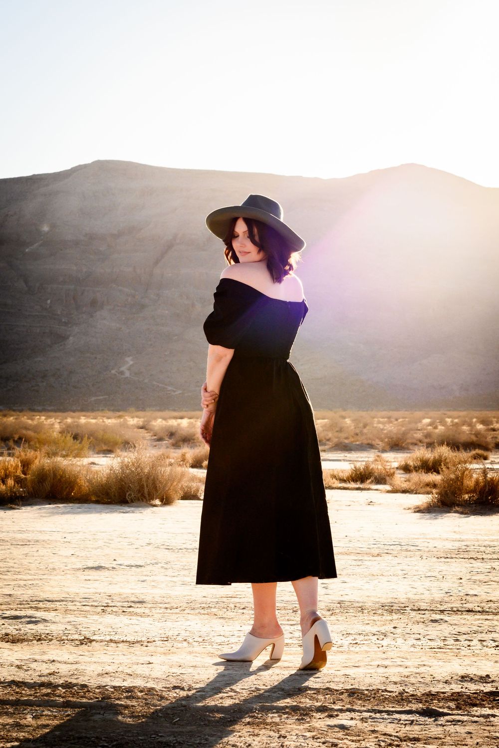 Woman in black off-shoulder dress and wide-brim hat poses in desert landscape at golden hour.