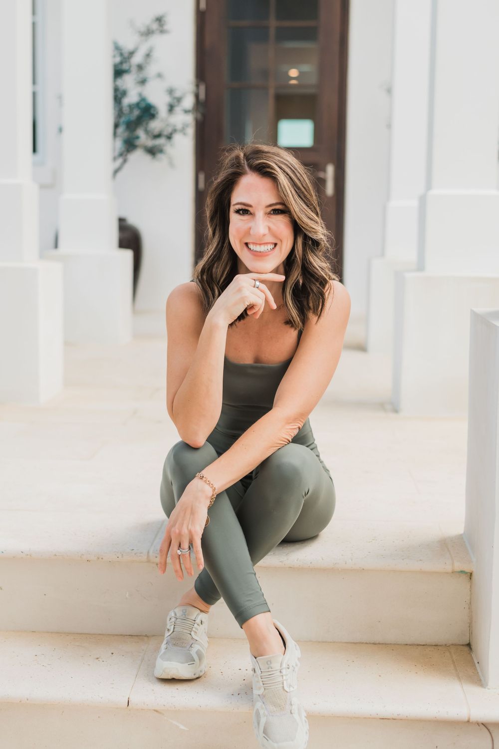 Woman in olive green athletic wear and sneakers sits smiling on white stairs in bright outdoor setting.