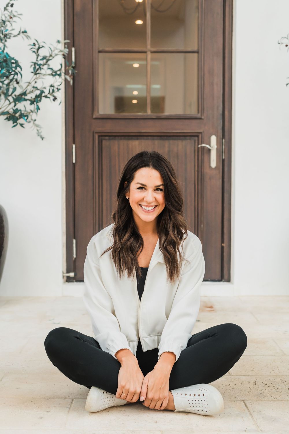 Casually dressed person sitting cross-legged on a floor in front of a wooden door with a light-colored outfit on.